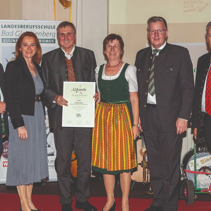 Fünf Erwachsene stehen auf einem roten Teppich und halten ein Zertifikat. Sie sind in formeller Kleidung gekleidet. Hinter ihnen steht ein Schild mit der Aufschrift 'Landesberufsschule Bad Goeggingen'. Im Hintergrund sind Musikinstrumente zu sehen.