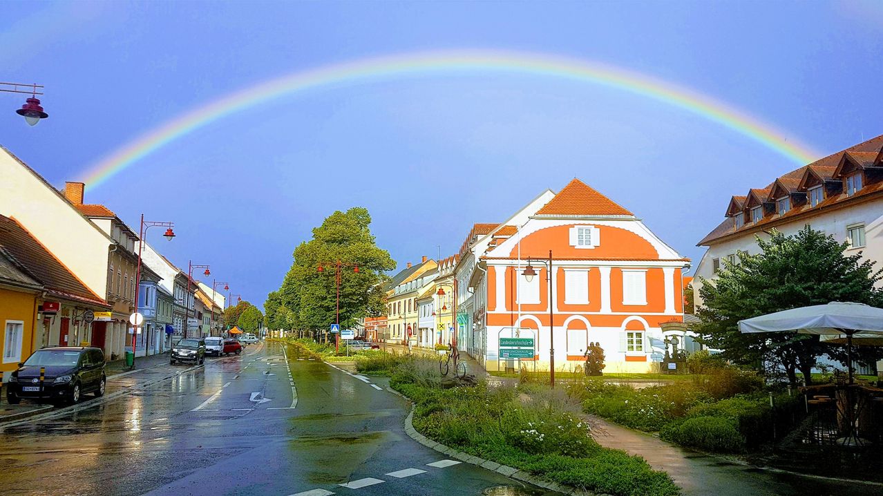 Ein Regenbogen wölbt sich über eine kleine Stadtstraße mit Gebäuden, geparkten Autos, Bäumen und Straßenlaternen. Der Himmel ist blau und es regnet.