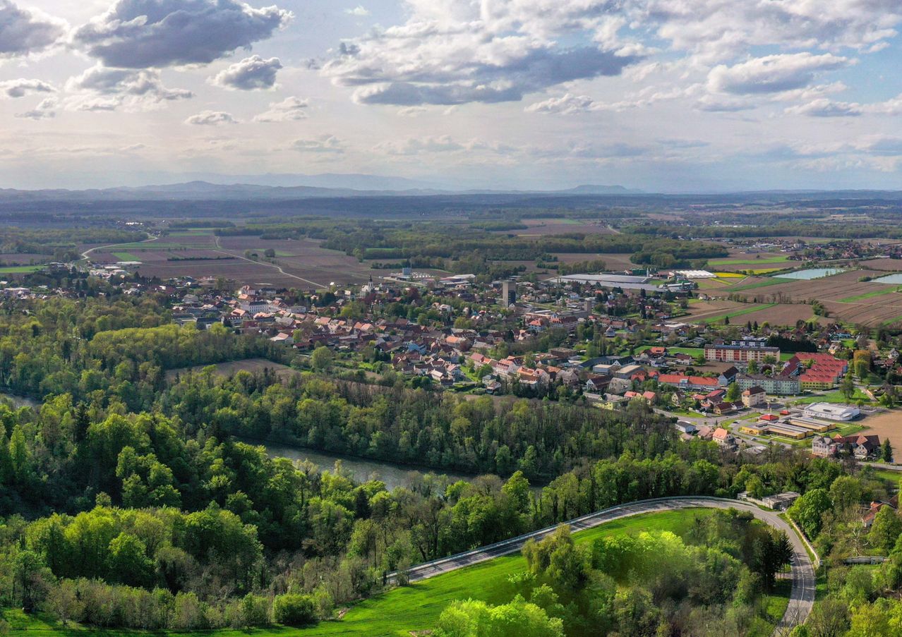 Luftaufnahme einer Stadt mit vielen Gebäuden und Häusern, umgeben von grünen Bäumen und einem Fluss unter einem bewölkten Himmel.