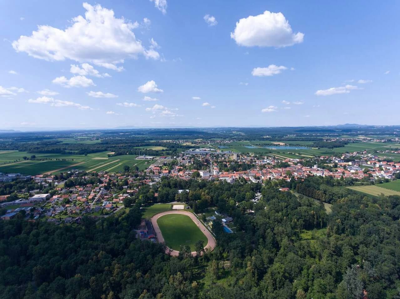 Luftaufnahme einer Stadt mit einem großen Feld und Stadion, umgeben von Bäumen und einem klaren blauen Himmel mit Wolken.
