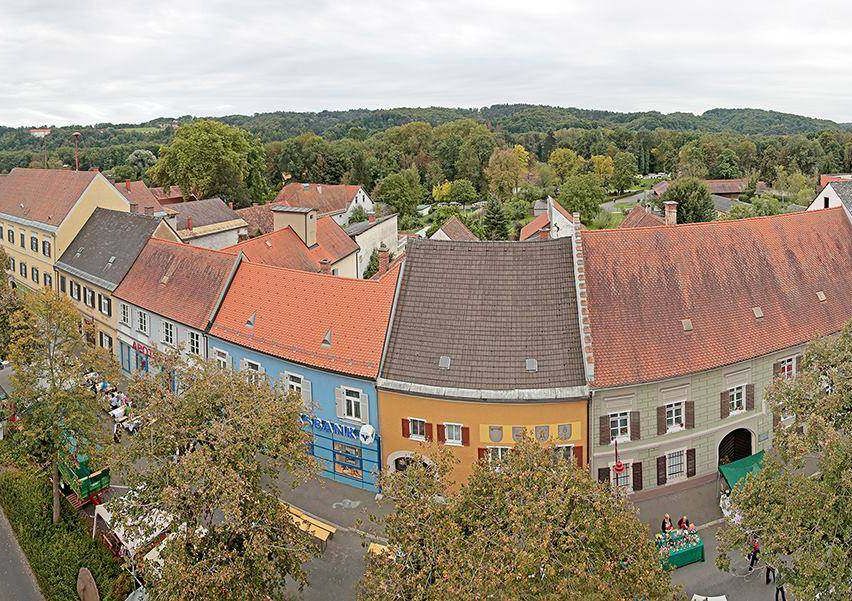 Luftaufnahme einer Stadt mit farbenfrohen Gebäuden, umgeben von Bäumen und einem bewölkten Himmel. Es gibt Leute, die auf der Straße gehen.