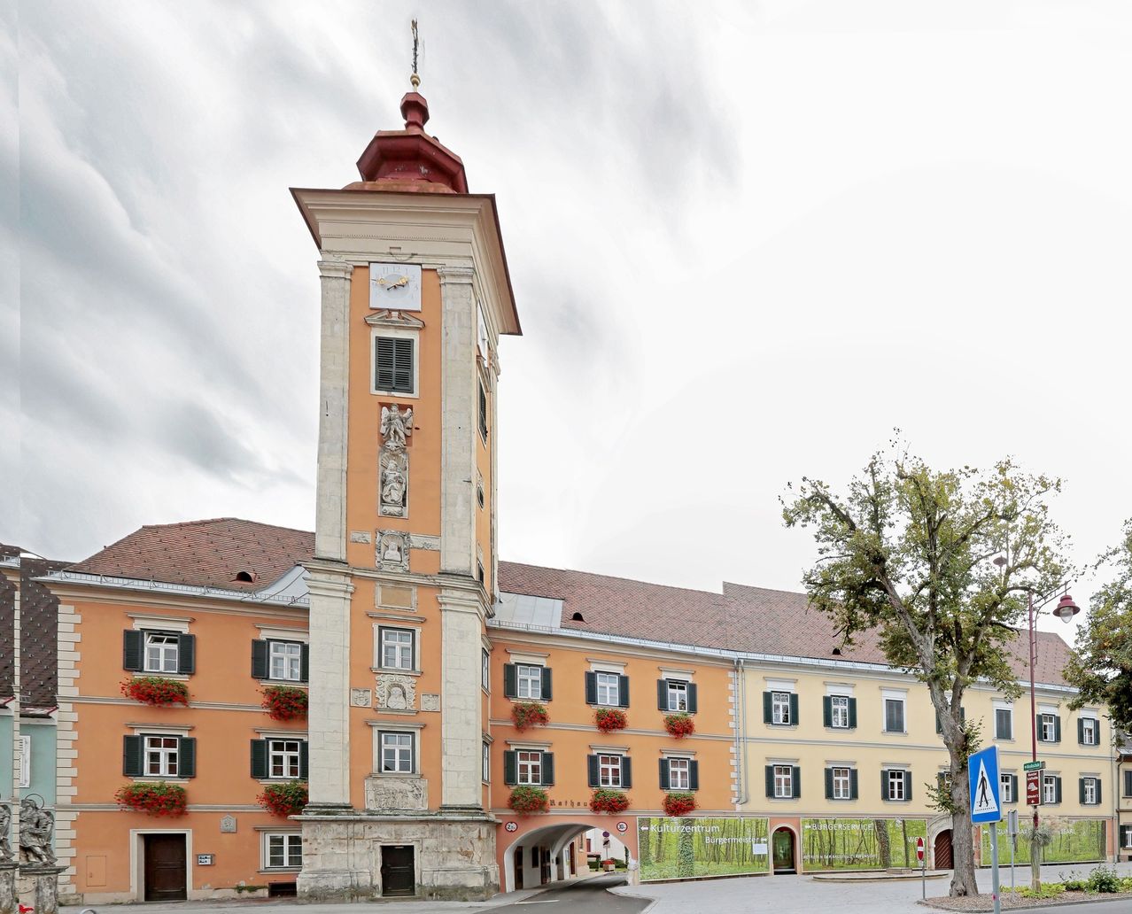 Ein Gebäude mit orangefarbener Außenseite und einem hohen Turm hat mehrere Fenster und dekorative Elemente. Der Turm zeigt eine Uhr und Statuen. Das Gebäude ist von Grünflächen und einem Baum umgeben.