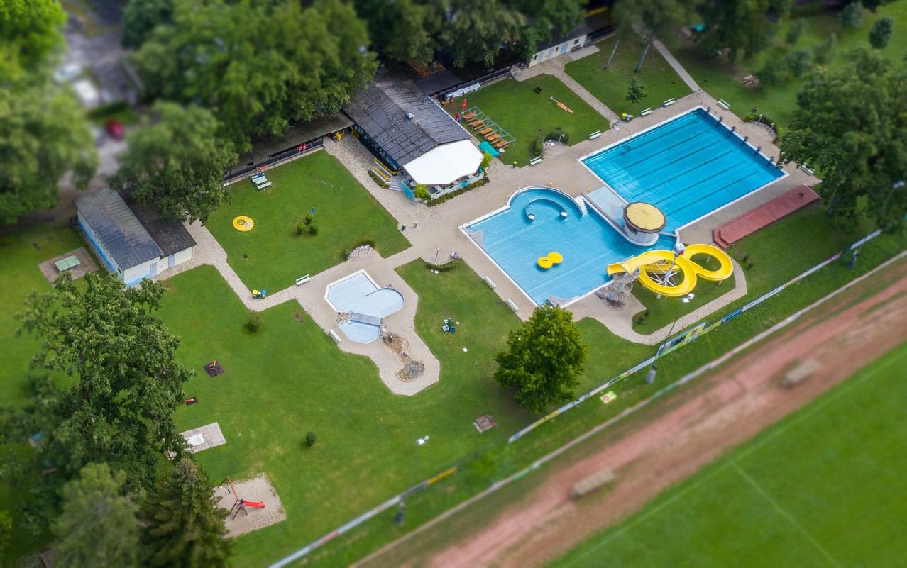 Ein Luftbild eines Freizeitparks mit einem großen Schwimmbad, Wasserrutschen und einem kleinen Pool mit Rutschen. Umgeben ist das Schwimmbad von einer Rasenfläche mit verschiedenen Spielgeräten.