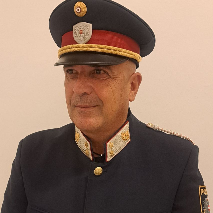 Ein älterer Mann in Uniform lächelt, trägt einen Hut mit Emblem und goldenen Akzenten. Die Uniform hat einen schwarzen und roten Kragen mit goldenen Sternen.