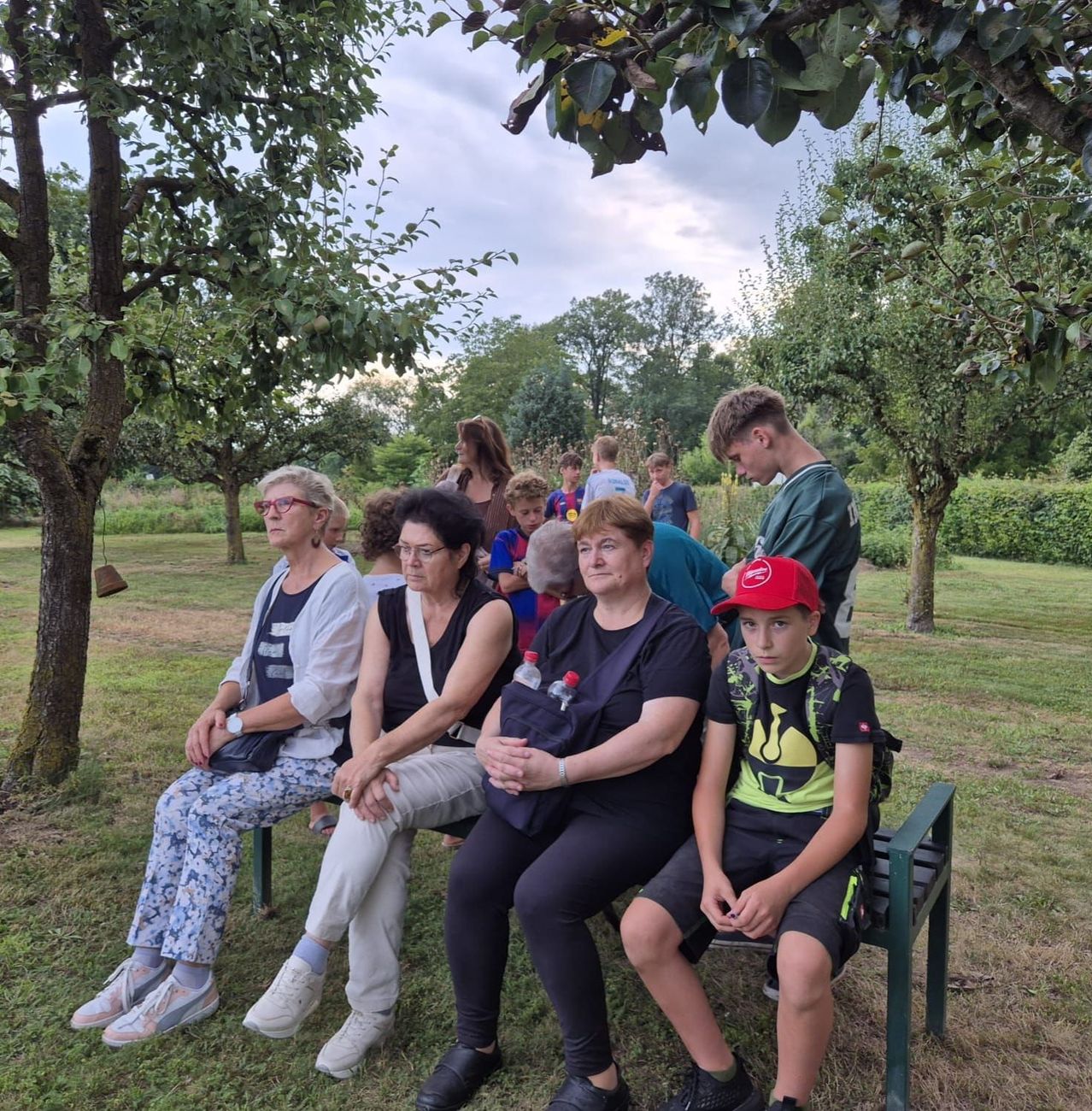 Eine Gruppe von Menschen, darunter Frauen und ein Junge, sitzt auf einer Bank in einem Park unter einem Baum. Einige tragen Brillen und haben Taschen dabei. Bäume und Gras sind im Hintergrund.