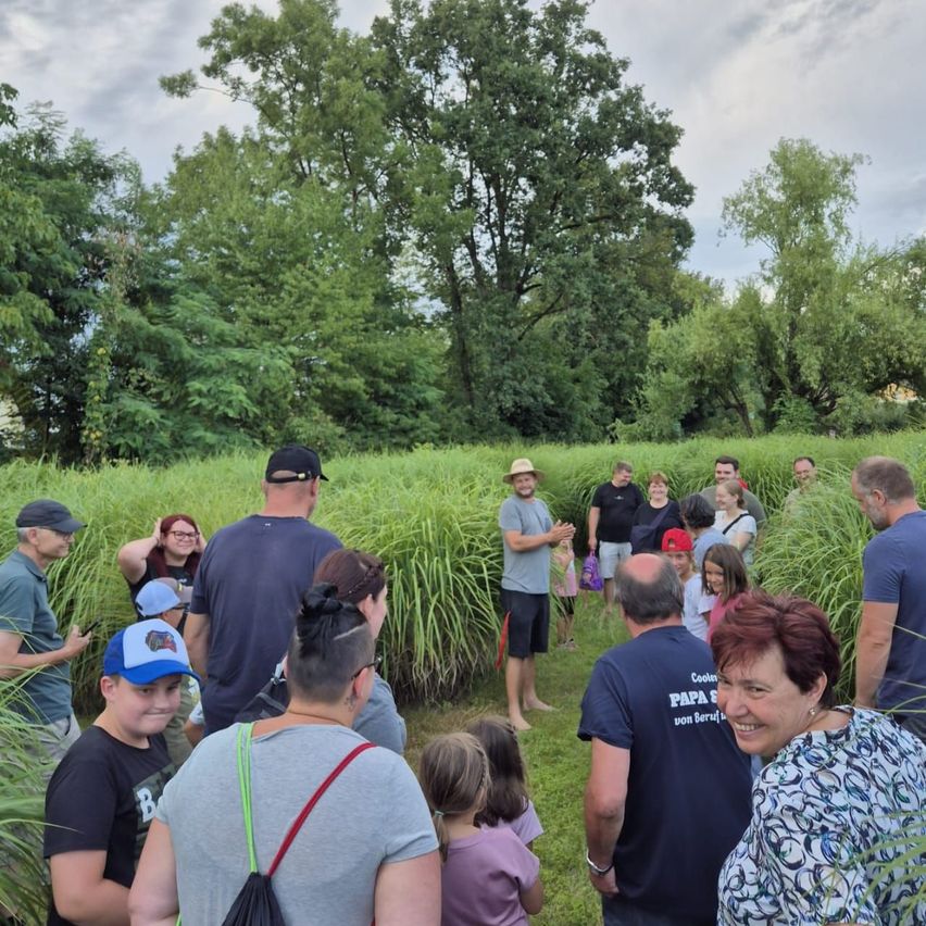 Eine Gruppe von Menschen, einschließlich Erwachsener und Kindern, steht in einem hohen Grasfeld mit Bäumen im Hintergrund. Einige tragen Hüte und Brillen. Ein Mann in einem braunen Hut spricht zu der Gruppe.