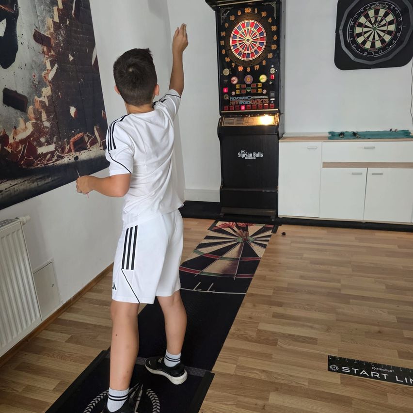 Ein junger Junge in einem weißen Sportanzug steht in einem Raum und wirft einen Dart auf ein Dartboard. Das Dartboard ist auf einem schwarzen Ständer montiert und ein Dart steckt bereits darin.