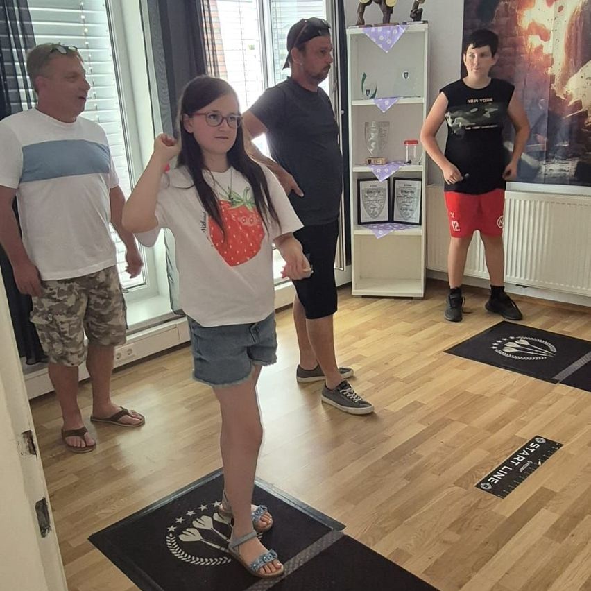 Vier Personen sind drinnen, zwei auf Dartbrettern, ein Mädchen mit Brille und Erdbeerhemd und ein Junge in roten Shorts. Dahinter ein Mann mit Cap und ein anderer Mann in einem weißen Shirt.