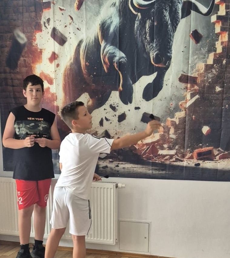 Zwei junge Jungen stehen vor einem großen Poster eines Stiers, der eine Mauer durchbricht. Ein Junge hält ein Mikrofon und scheint zu sprechen.