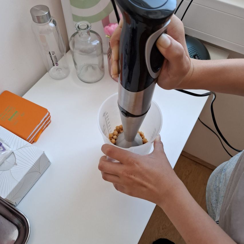 Eine Person benutzt einen Stabmixer, um Bohnen in einem Plastikbecher auf einem weißen Tisch zu pürieren.