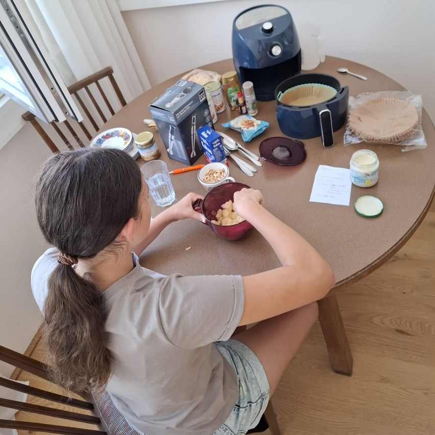 Ein junges Mädchen sitzt an einem Tisch mit einer Schüssel Essen, umgeben von verschiedenen Küchenutensilien. Es gibt einen Kuchenboden, ein Glas Erdnussbutter und eine Box eines Lebensmittelprozessors.