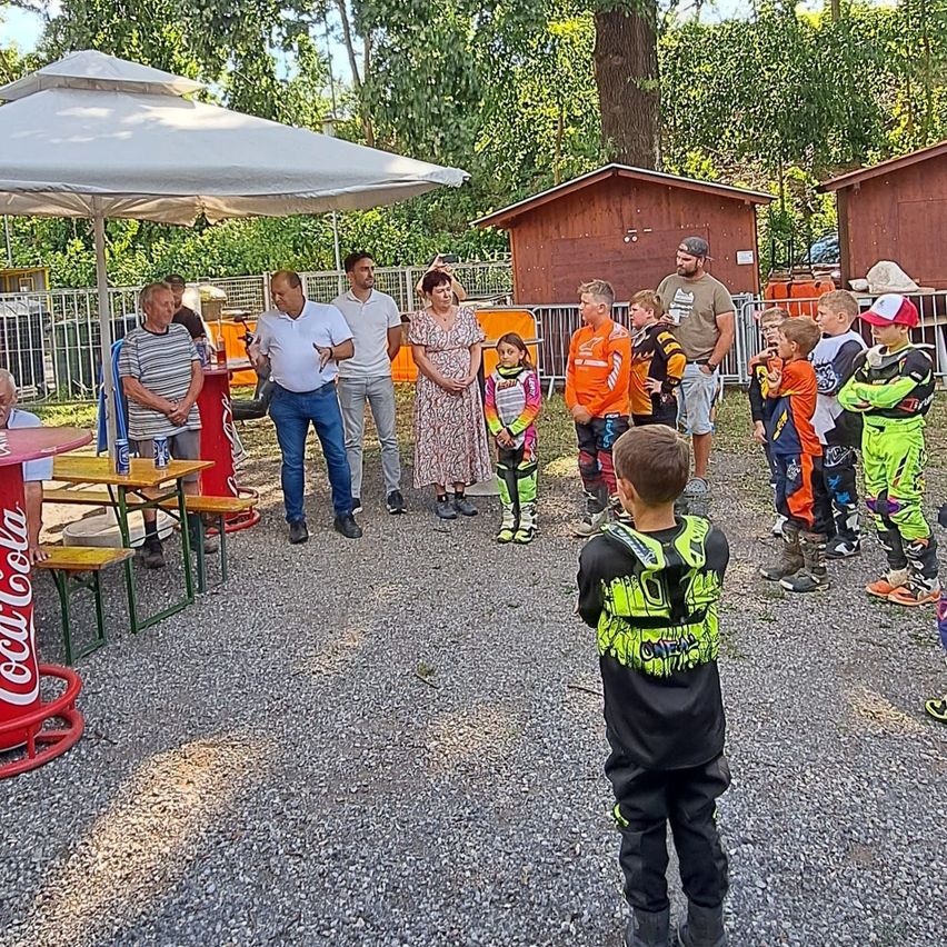 Eine Gruppe von Menschen, einschließlich Kinder in Rennanzügen, versammeln sich in einem Außenbereich mit einem Coca-Cola-Tisch und einem Schirm.