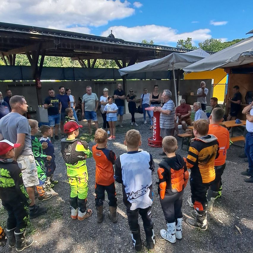 Eine Gruppe von Kindern und Erwachsenen versammelt sich in einem Außenbereich, wahrscheinlich für eine Sportveranstaltung. Die Kinder tragen Sportausrüstung und hören einem Erwachsenen zu, der einen roten Becher hält. Hinter ihnen stehen einige Zelte und ein Gebäude.
