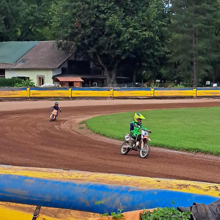Zwei Personen fahren auf einer Dirt-Rennstrecke mit Dirtbikes, eine in grüner Kleidung und Helm, die andere in Schwarz. Ein Haus mit grünem Dach ist im Hintergrund zu sehen.