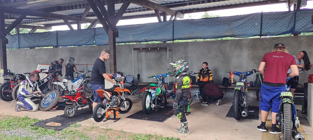 Ein Mann justiert ein Motocross-Motorrad, während ein Junge in einem Helm zusieht. Ein weiteres Motorrad steht auf einem Ständer und ein drittes Motorrad ist im Hintergrund.