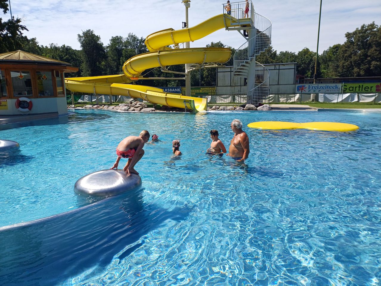 Eine Gruppe von Menschen schwimmt in einem großen Pool mit einer gelben Wasserrutsche im Hintergrund. Eine Person steht auf einem silbernen Schwimmkörper.