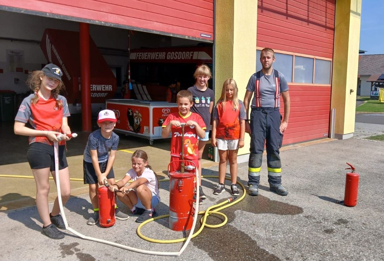 Eine Gruppe von Kindern und zwei Erwachsenen steht vor einer Feuerwehrwache und hält Feuerlöscher und eine Feuerwehrschläuche. Die Kinder tragen Uniformen und scheinen an einer Brandschutzvorführung teilzunehmen.