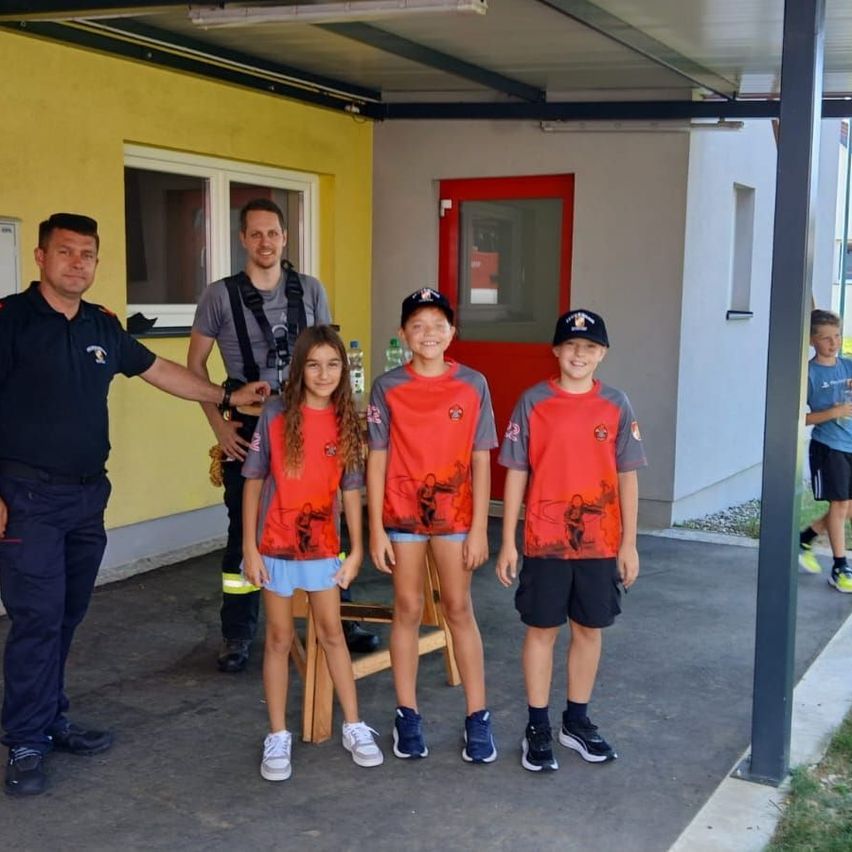 Drei Kinder in passenden roten und grauen Uniformen stehen neben zwei Männern, einer in blauer Uniform und der andere in einem grauen Shirt, vor einem gelben Gebäude.