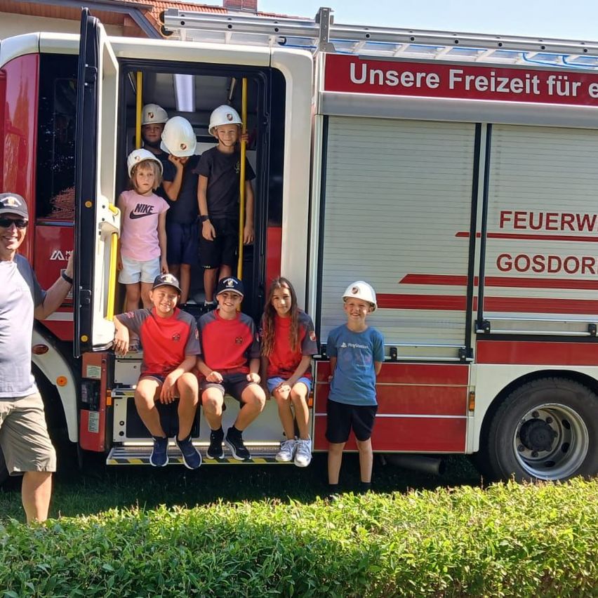 Eine Gruppe von Kindern und einem Mann stehen vor einem Feuerwehrauto. Die Kinder tragen Helme und rote Hemden und sitzen im Inneren des Autos. Der Mann trägt eine Sonnenbrille und eine Uhr.
