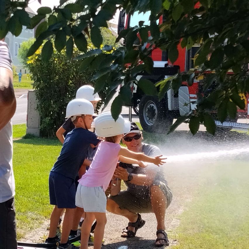 Drei Kinder mit weißen Helmen stehen auf einem Rasen. Ein Mann mit Sonnenbrille und Uhr hält einen Wasserschlauch und spritzt Wasser auf die Kinder. Ein Feuerwehrauto ist im Hintergrund.