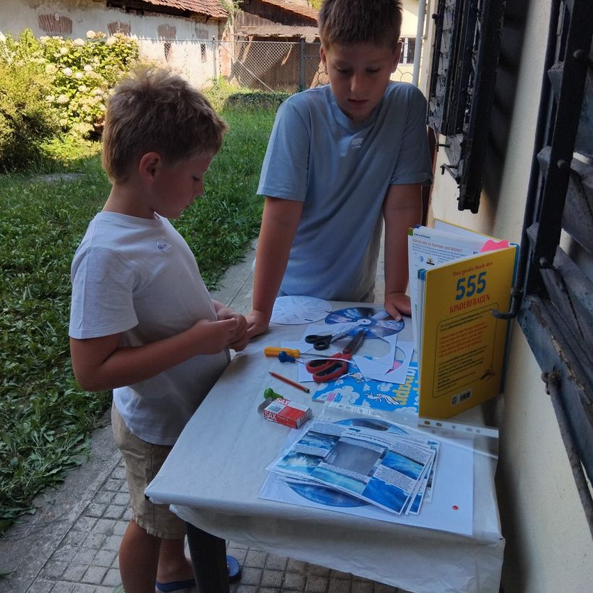 Zwei Jungen stehen an einem Tisch mit Bastelmaterialien, einer schaut nach unten, während der andere ein Buch mit dem Titel '555 Kindertagen' ansieht.