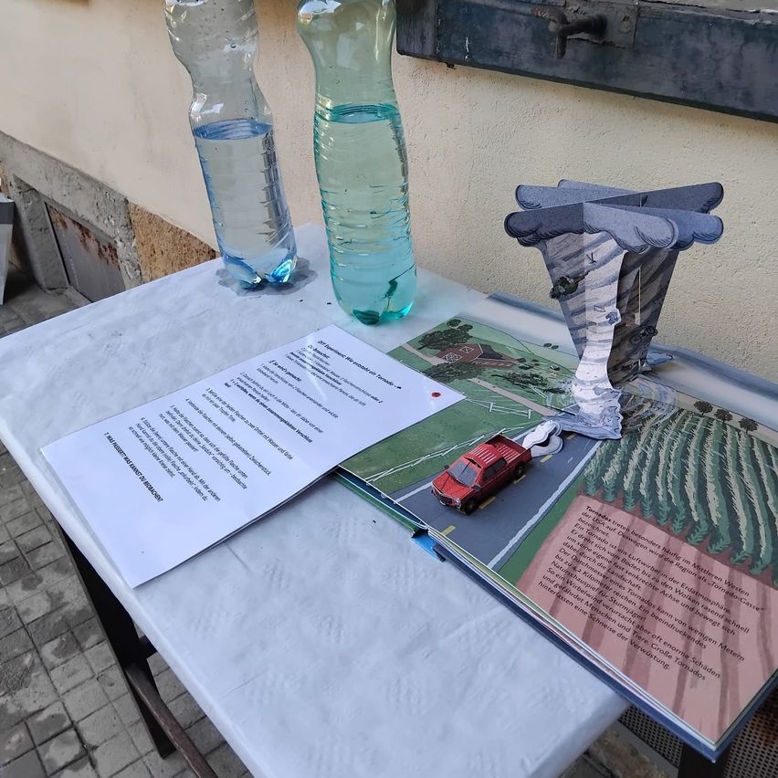 Ein Tisch mit zwei klaren Wasserflaschen, einem Papier mit Text und einem offenen Buch mit einem roten Spielzeugauto und einem Papierbaum darauf.