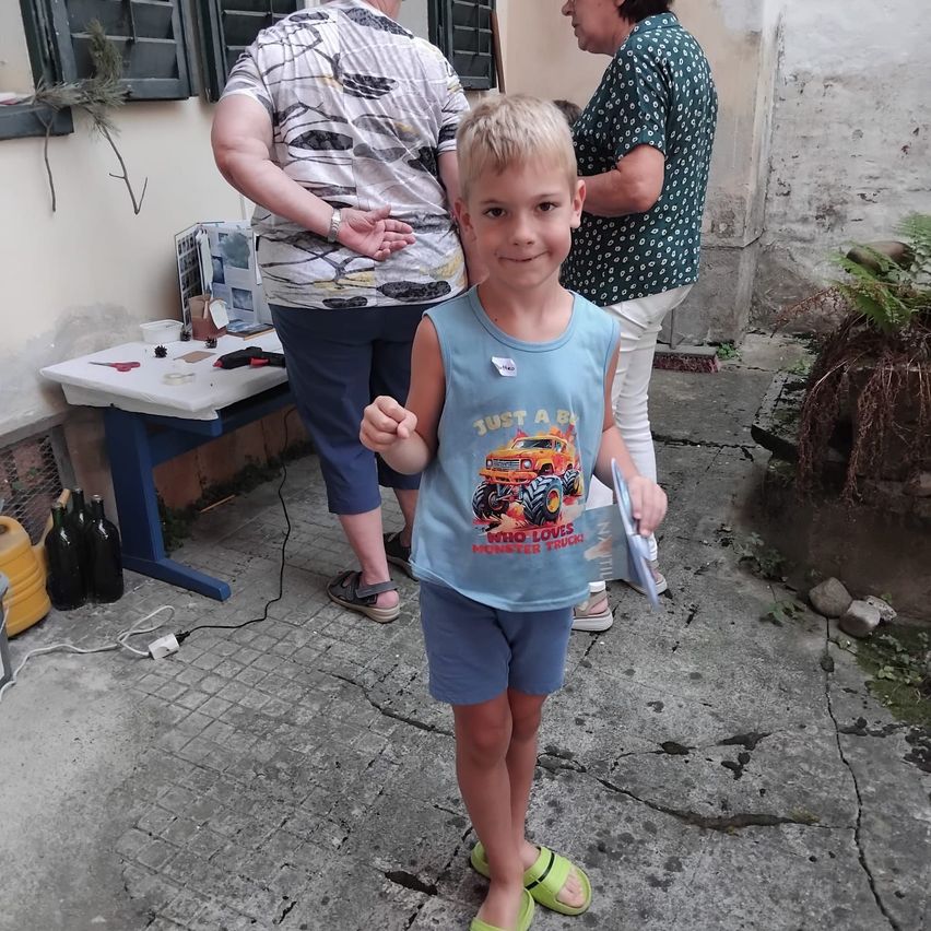 Ein junger Junge mit einem blauen T-Shirt, auf dem 'Nur ein Junge, der Monster Trucks liebt' steht, steht vor einem Tisch, hinter dem eine Frau steht.