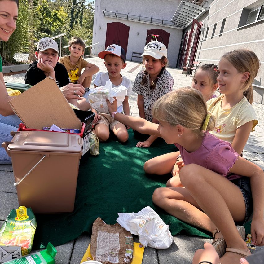 A group of children and an adult are sitting on the floor around a box of snacks. The adult is handing out snacks to the children.