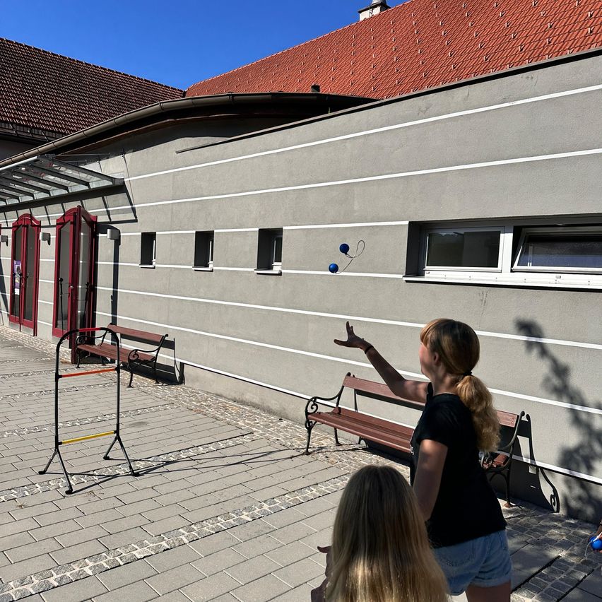 A woman practices juggling blue balls outside a building with a red roof and gray walls. Two benches and a metal rack are nearby.