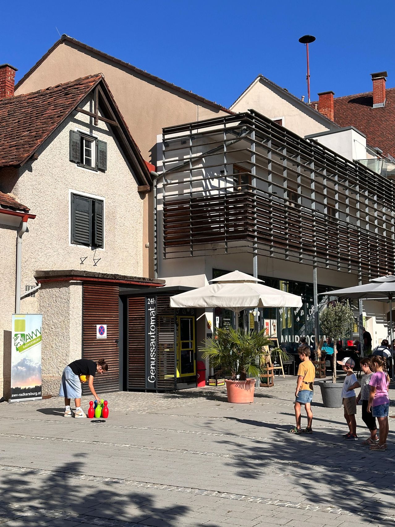 A woman is playing a game of bocce ball on a sunny day in front of a building with a sign that reads 'Genussautomat'. Several children are watching her. There are potted plants and umbrellas in the area.