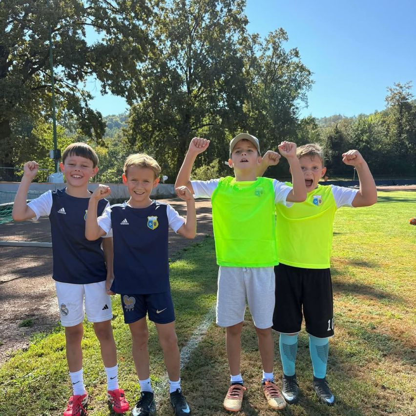 Four young boys in soccer uniforms stand on a field, flexing their muscles and smiling for the camera. They are wearing different colored jerseys and shorts, and the background shows a grassy field with trees.