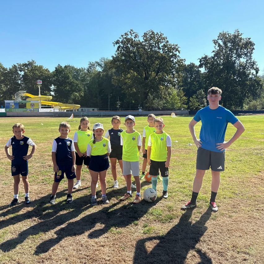 A group of young children in yellow and blue uniforms are standing on a grassy field with a soccer ball in front of them, and a young man in a blue shirt is standing behind them.