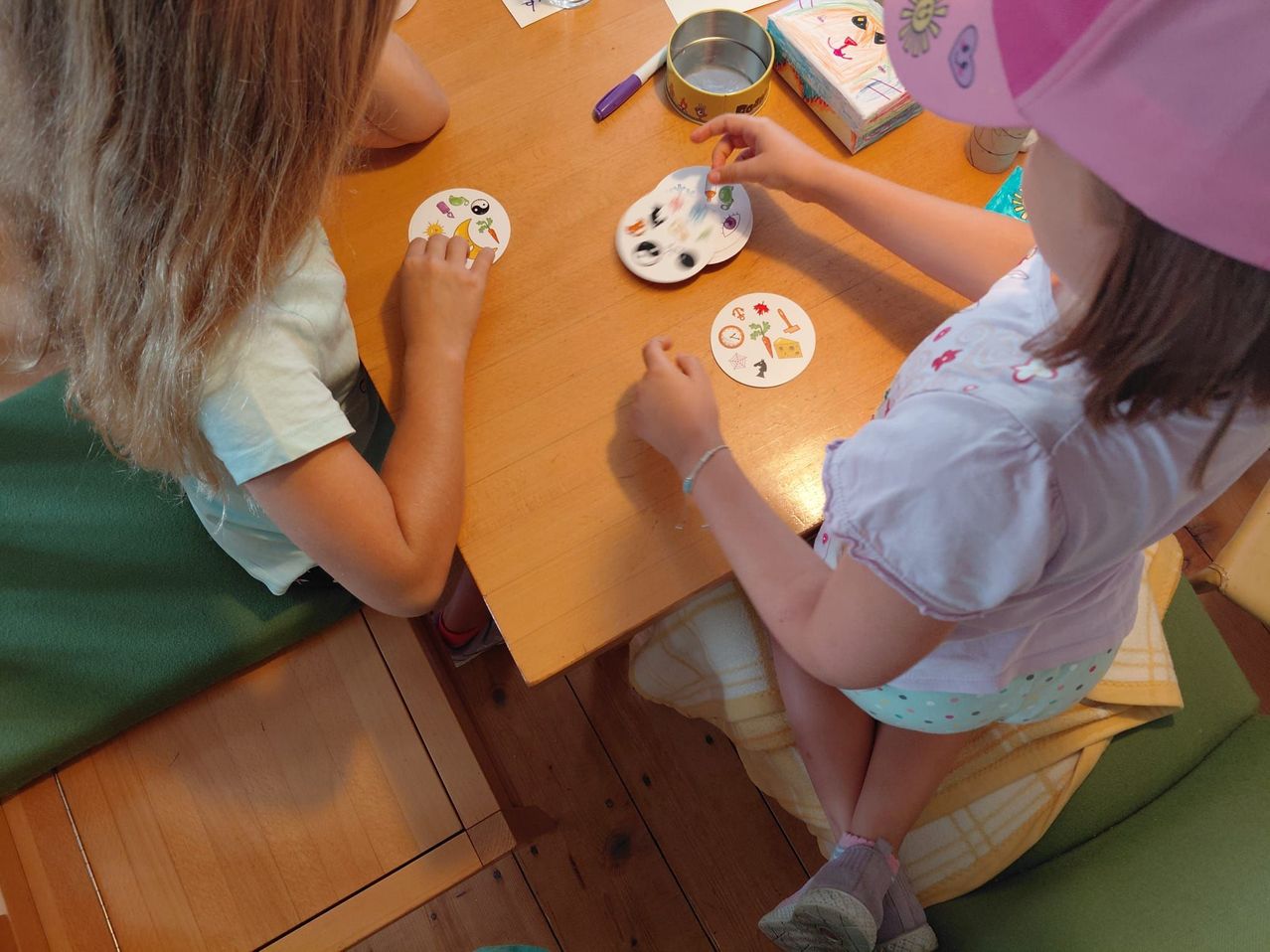 Two young girls are sitting at a wooden table, engaged in an activity. One girl is holding a circular sticker with various objects on it, while the other is holding a similar sticker. They are both wearing bracelets and appear to be focused on their task.