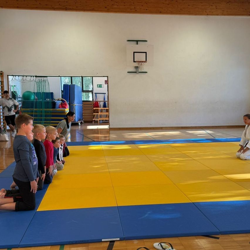 A group of children and an adult are kneeling on a blue and yellow mat in a gym. A person in a white uniform is sitting on the mat.