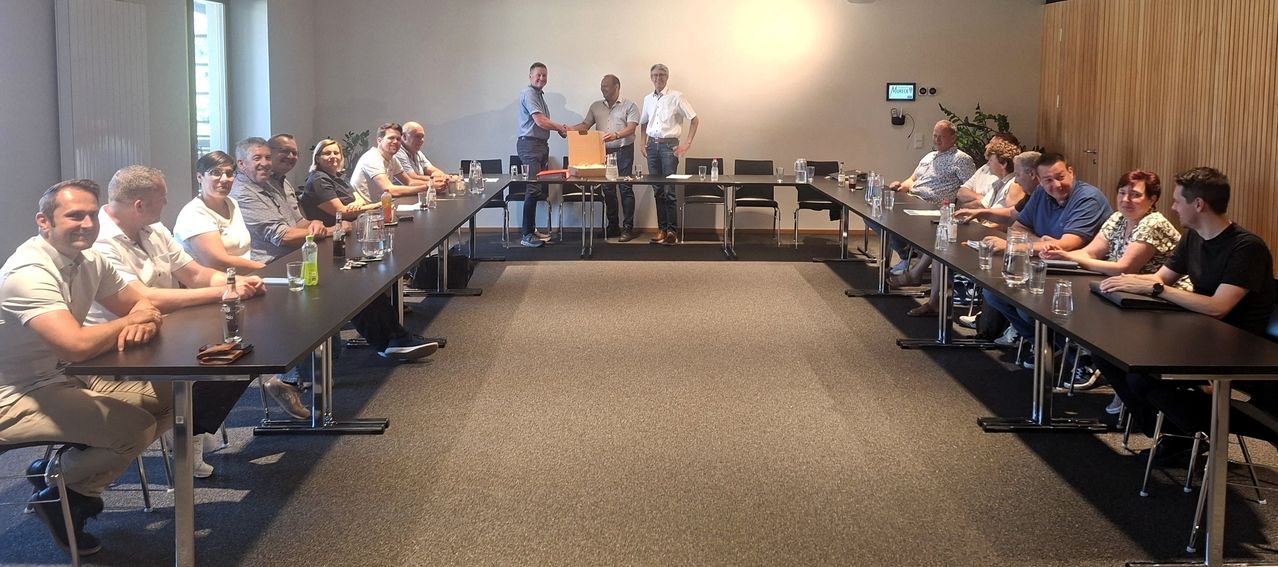 Eine Gruppe von Männern versammelt sich um einen Tisch. Zwei Männer stehen in der Mitte und schütteln Hände, während die anderen sitzen. Gläser und Flaschen stehen auf dem Tisch.