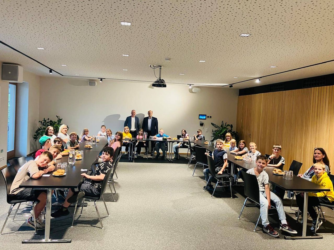 Eine Gruppe von Kindern sitzt in einem Klassenzimmer an Tischen. Zwei Männer stehen in der Mitte. Sie alle schauen nach vorne und scheinen an einer Aktivität teilzunehmen.