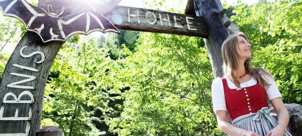 Eine Frau in traditioneller Kleidung steht unter einem hölzernen Schild mit einer Fledermaus-Design und dem Wort Hohle, umgeben von üppiger Grün.