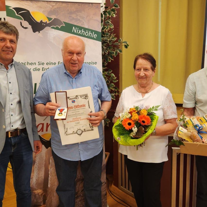 Ein Mann und eine Frau halten einen Blumenstrauß und ein Zertifikat. Der Mann hält eine Medaille in der anderen Hand. Hinter ihnen ist ein Banner mit einem Bild einer Fledermaus.