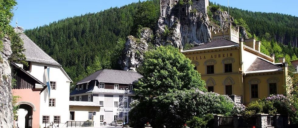 Eine kleine Stadt mit einer Burg auf einem felsigen Felsvorsprung darüber, umgeben von Bäumen. Ein gelbes Gebäude mit 'VILLA AUHEI' darauf.