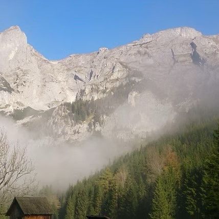 Ein nebliger Bergrücken mit einer kleinen Hütte im Vordergrund. Der Himmel ist klar und blau.