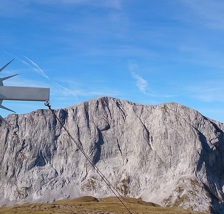 Ein großes Kreuz mit einem Kruzifix steht auf einem Berggipfel, umgeben von einer Bergkette unter einem blauen Himmel.