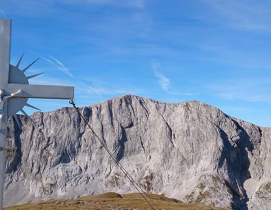 Ein großes Kreuz mit einem Kruzifix steht auf einem Berggipfel, umgeben von einer Bergkette unter einem blauen Himmel.