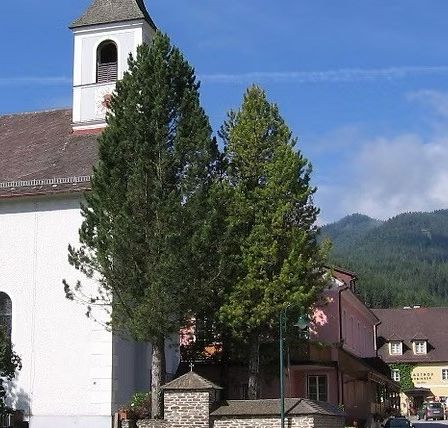 Eine weiße Kirche mit braunem Dach und einem Kirchturm, mit Bäumen davor und Bergen im Hintergrund. Es gibt auch Häuser und ein Auto in der Szene.