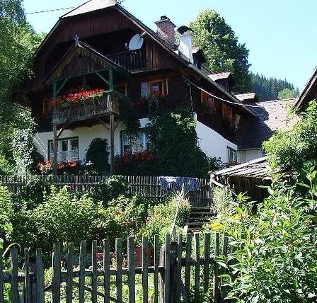 Ein traditionelles Holzhaus mit Balkon und Blumenkästen, umgeben von einem Garten und einem Holzzaun, in einem ländlichen Gebiet.