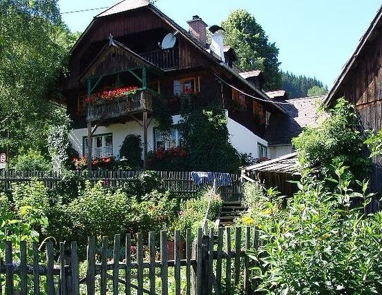 Ein traditionelles Holzhaus mit Balkon und Blumenkästen, umgeben von einem Garten und einem Holzzaun, in einem ländlichen Gebiet.