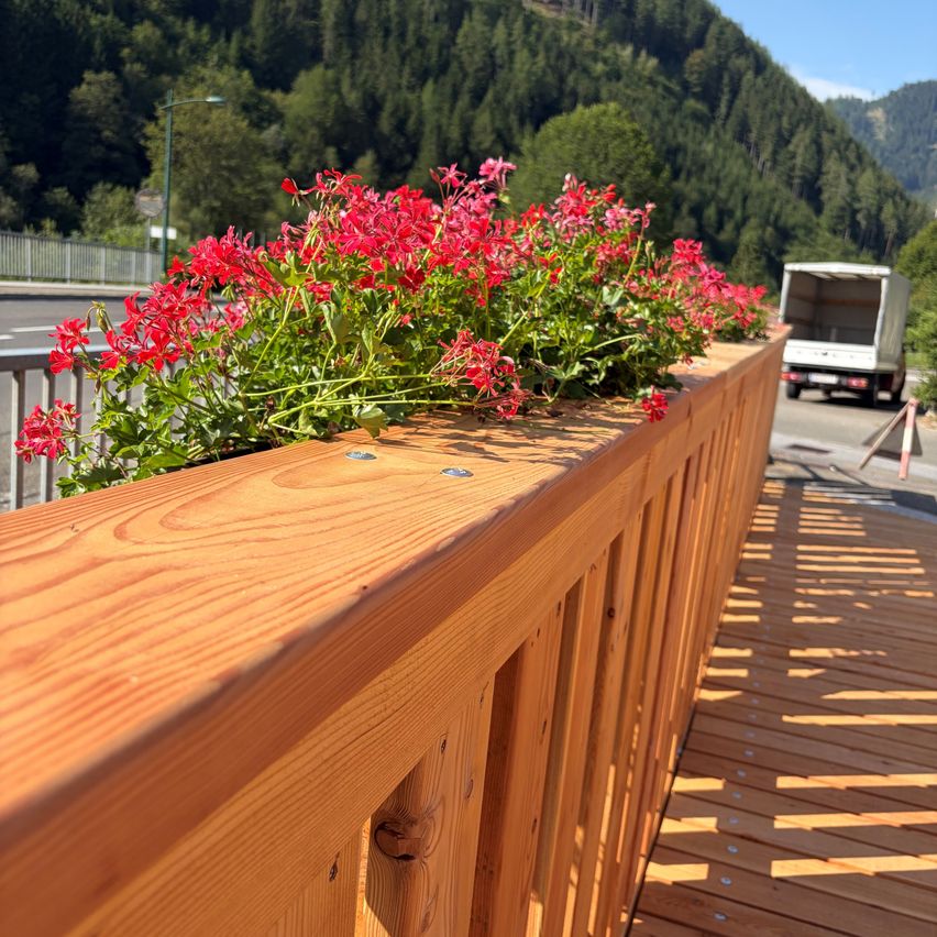 A wooden balcony railing with red flowers in a planter, overlooking a road with a truck and mountains in the background.