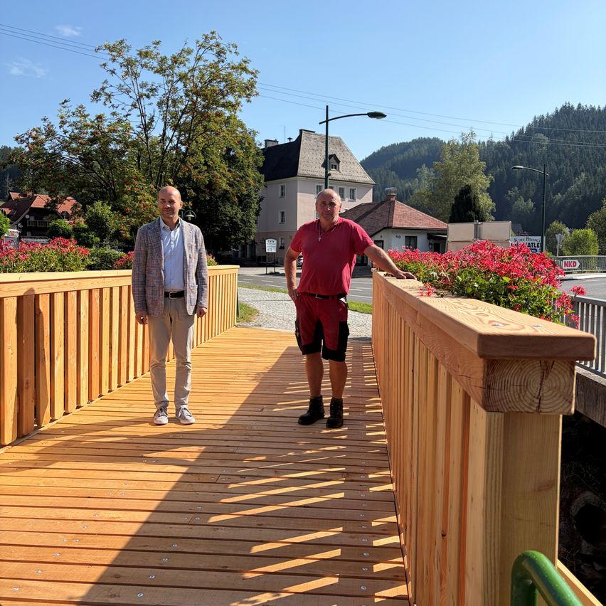 Two men stand on a wooden bridge with railings, one wearing a red shirt and the other in a gray jacket. Behind them, a building with a red roof and a tree with red flowers is visible.