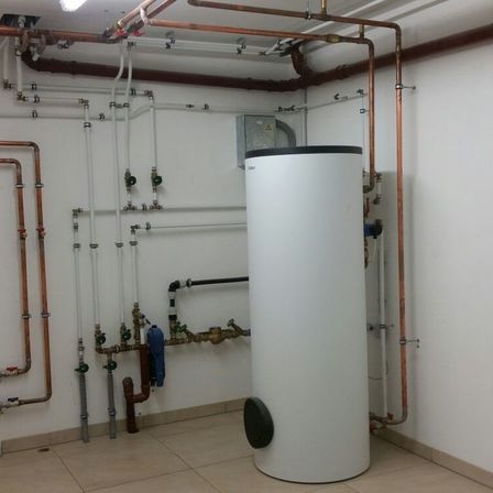 Ein großer weißer Wasserspeicher ist an der Wand montiert, umgeben von Kupfer- und weißen Rohren. Der Raum hat weiße Wände und einen gefliesten Boden.