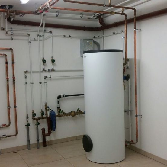 Ein großer weißer Wasserspeicher ist an der Wand montiert, umgeben von Kupfer- und weißen Rohren. Der Raum hat weiße Wände und einen gefliesten Boden.