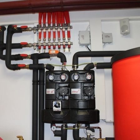 Ein Nahaufnahme einer roten und schwarzen Wasserheizungsanlage, die an einer weißen Wand mit verschiedenen Rohren und Messgeräten montiert ist.